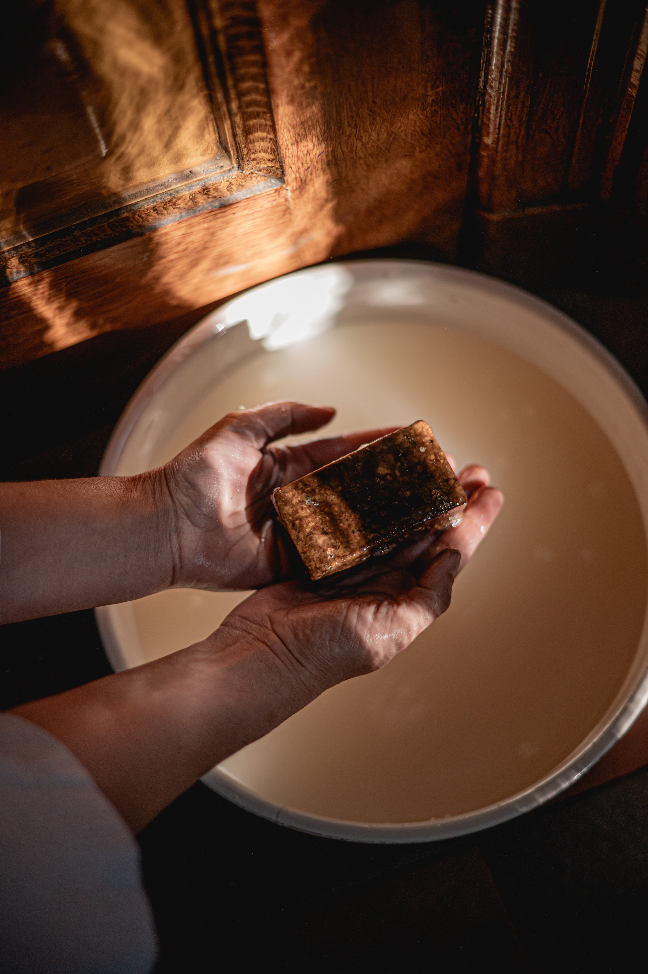 warm sfeerbeeld van twee handen die boven een emaille bak water een schuimend blok african black soap van Namashea vast houdt